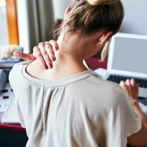 A person seen from behind, clutching their neck and shoulder in pain while working on a laptop.
