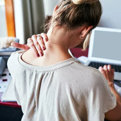 A person seen from behind, clutching their neck and shoulder in pain while working on a laptop.