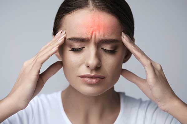 A woman with her eyes closed and furrowed brows, pressing her hands to her temples, with a red glow on her forehead indicating a severe headache or migraine.