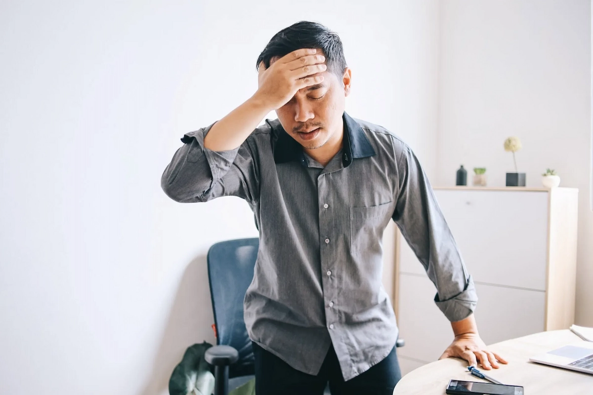 A man in a grey shirt stands with his hand on his forehead, looking distressed and tired in an office setting.