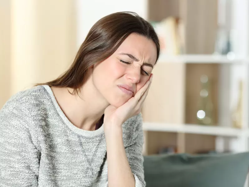 A young woman with long brown hair, wearing a grey sweater, presses her hand to her right cheek with a pained expression, eyes closed and brows furrowed, indicating a toothache or facial pain.