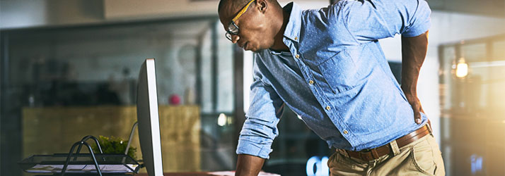 A man in glasses and a blue shirt holds his lower back in an office, indicating back pain while standing near a computer monitor.
