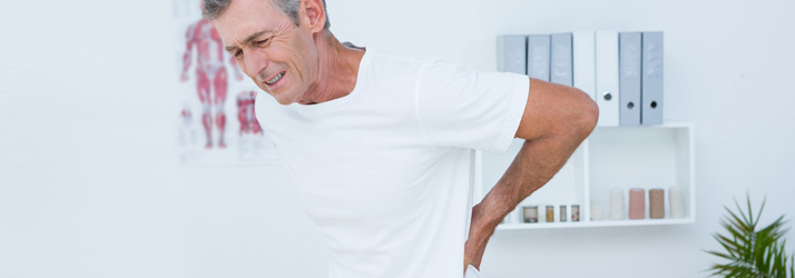 A man in a white t-shirt grimaces in pain while holding his lower back with both hands in a doctor's office.