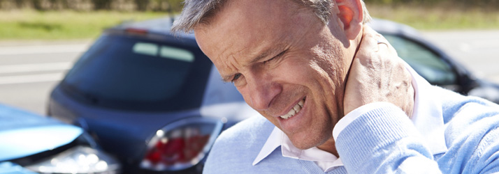 A man grimacing in pain and holding his neck, with two damaged cars visible in the blurred background, indicating a car accident.