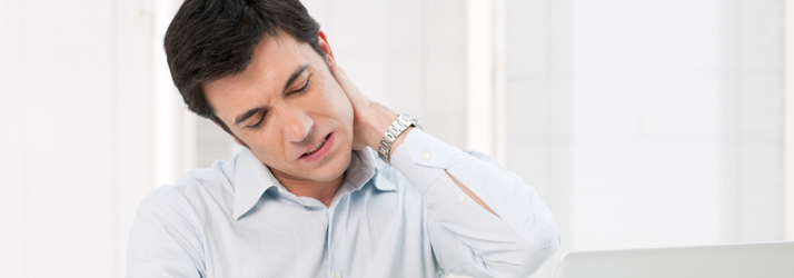 Man with dark hair, wearing a light blue shirt, grimacing and holding the back of his neck with his left hand due to pain.