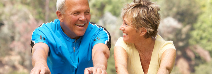 A smiling older man in a blue shirt and an older woman in a yellow top look at each other while cycling outdoors.