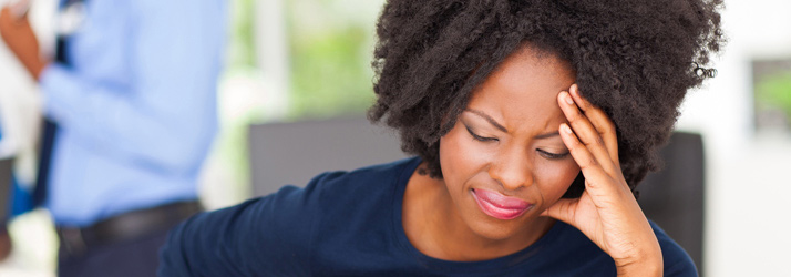Close-up of a Black woman with curly hair, eyes closed and hand pressed to her temple, indicating a headache or stress.
