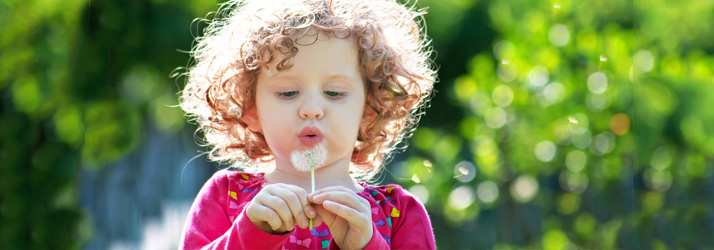 A young child with curly hair blows on a dandelion, scattering its seeds in a sunny, green outdoor setting.