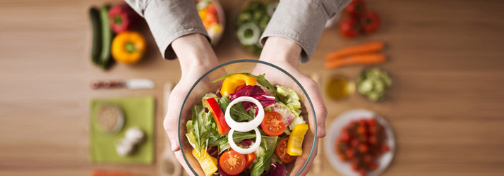 Overhead view of hands holding a glass bowl of colorful salad with mixed greens, tomatoes, bell peppers, and onion rings, surrounded by blurred fresh vegetables on a wooden table.