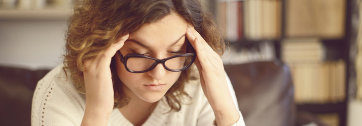 A young woman with curly brown hair and glasses, pressing her hands to her temples, looking down with a pained or stressed expression.