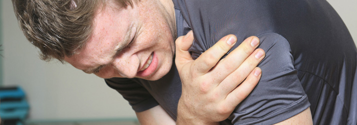 A man grimacing intensely in pain, clutching his right shoulder with his left hand. His eyes are shut and teeth clenched, indicating severe discomfort.