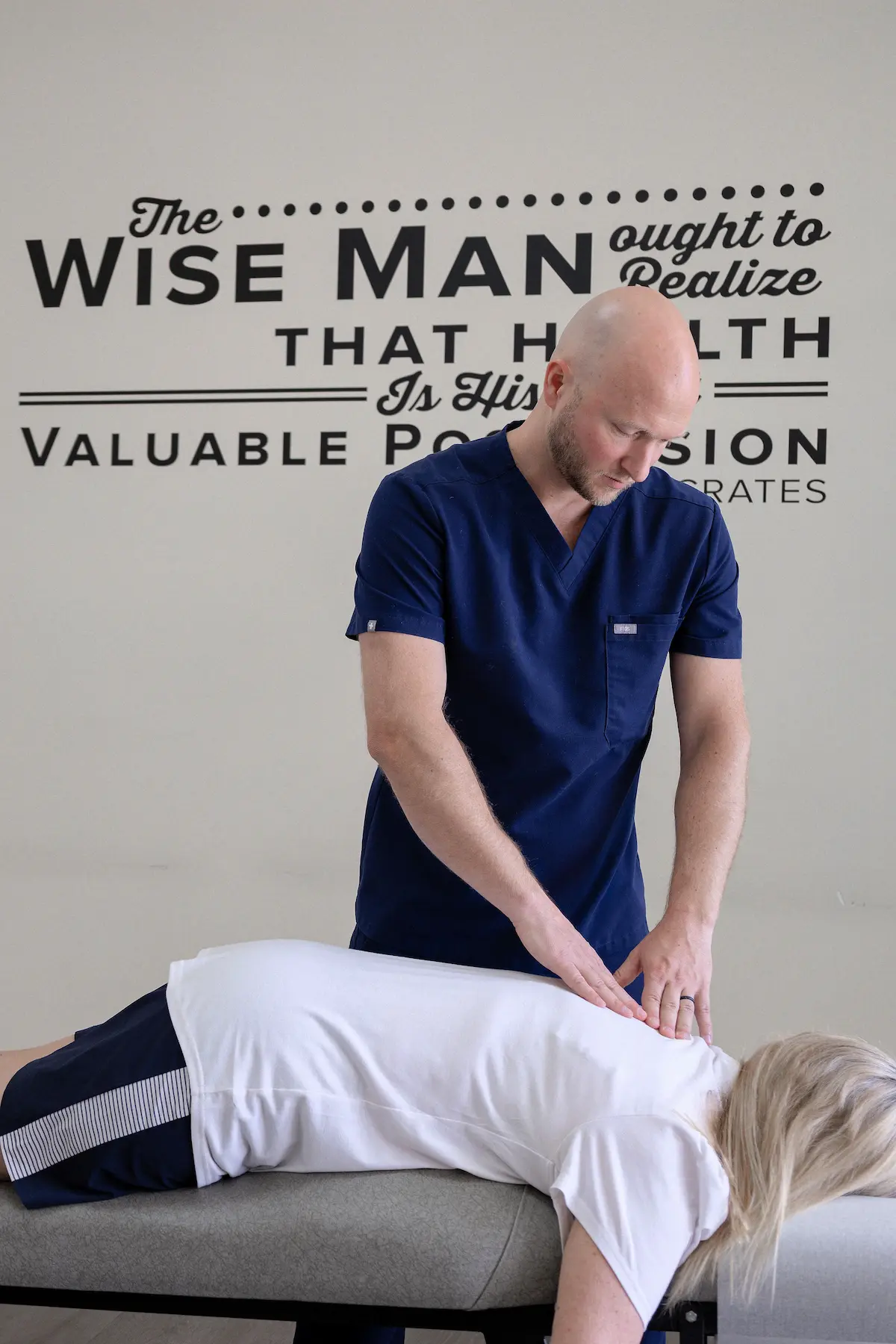 A bald man in blue scrubs massages the upper back of a person lying face down on a table. A quote about health is on the wall.