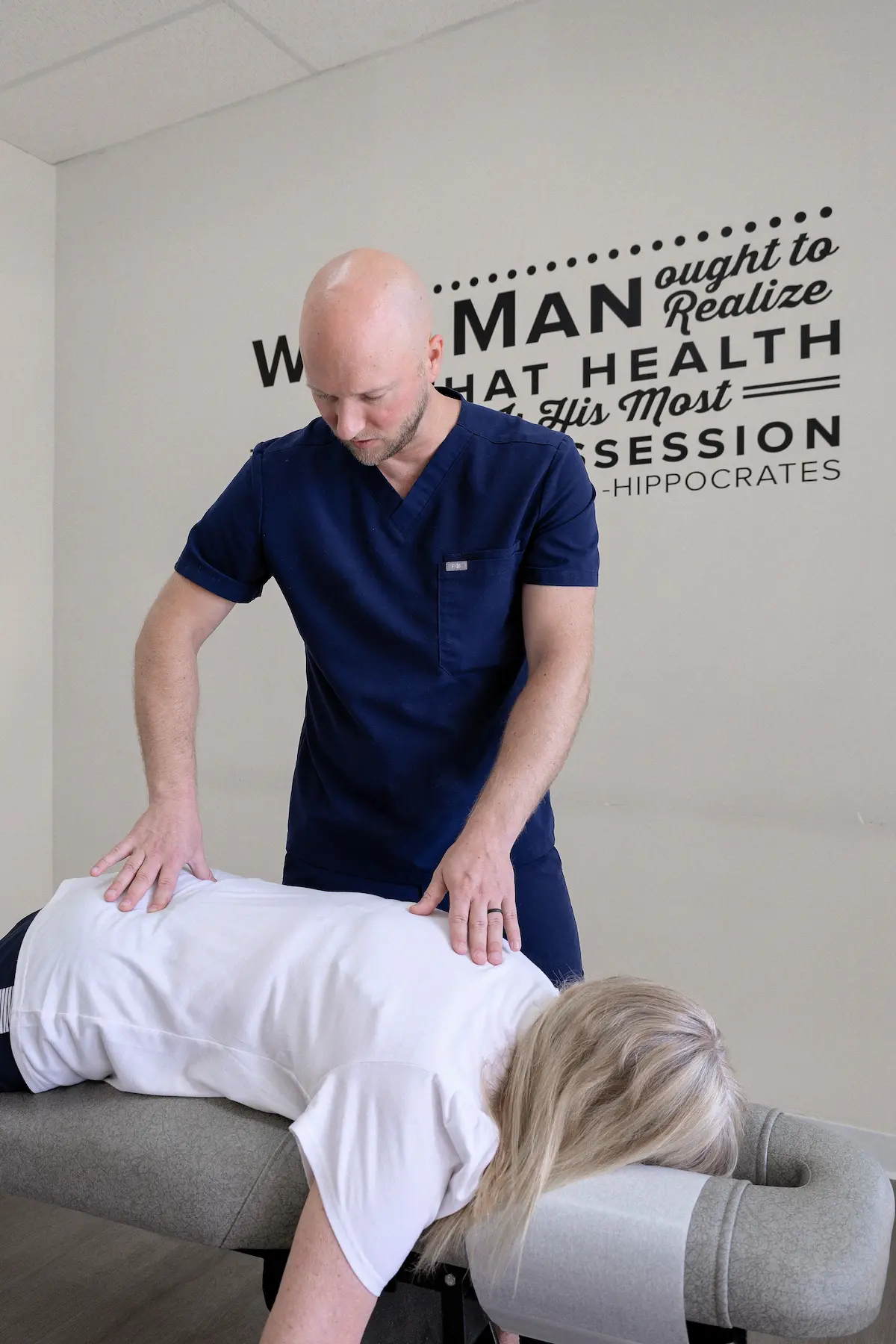 A male chiropractor in blue scrubs adjusts the back of a female patient lying face down on a chiropractic table.