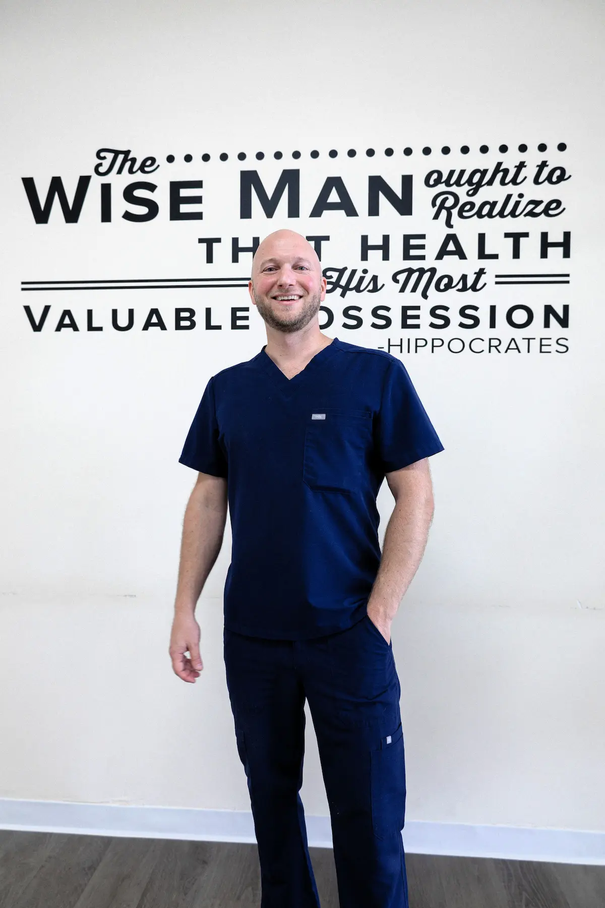A smiling bald man in navy blue scrubs stands in front of a white wall with a black quote decal by Hippocrates about health.